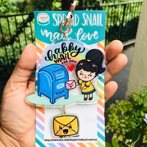 Pu&ograve; includere: Un portachiavi in acrilico colorato con una ragazza dei cartoni animati che consegna una lettera a una cassetta delle lettere blu con il testo "Happy Mail for You!" e un portachiavi a forma di busta gialla pi&ugrave; piccolo con un viso sorridente.