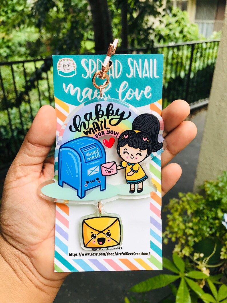 Pu&ograve; includere: Un portachiavi in acrilico colorato con una ragazza dei cartoni animati che consegna una lettera a una cassetta delle lettere blu con il testo "Happy Mail for You!" e un portachiavi a forma di busta gialla pi&ugrave; piccolo con un viso sorridente.