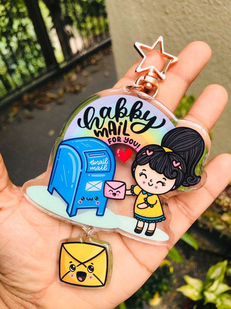 Pu&ograve; includere: Un portachiavi in acrilico trasparente con un design di cassetta postale blu con un viso sorridente e la scritta "snail mail". Una ragazza dei cartoni animati con una coda di cavallo tiene una busta rosa e sorride. Il testo "happy mail for you!" &egrave; scritto in corsivo nero. Il portachiavi ha un ciondolo a forma di stella dorata.
