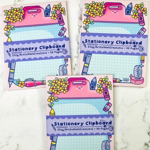 May include: Three rectangular stationery pads featuring a pink clipboard illustration with a grid paper center. The pads are wrapped in a purple band labeled Stationery Clipboard Etsy/ArtfulGeetCreations 50 Pages, decorated with colorful floral and school supply graphics.
