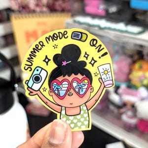 May include: A yellow sticker with a cartoon illustration of a person wearing heart-shaped sunglasses with a beach scene. The sticker has the text "SUMMER MODE ON!" and the person is holding a camera and sunscreen.