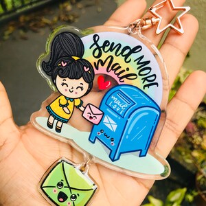 Pu&ograve; includere: Un portachiavi in acrilico colorato con una ragazza dei cartoni animati che tiene una busta rosa e una cassetta delle lettere blu con il testo "Send More Mail" e un arcobaleno. Il portachiavi include anche un ciondolo a forma di busta verde con un viso felice.