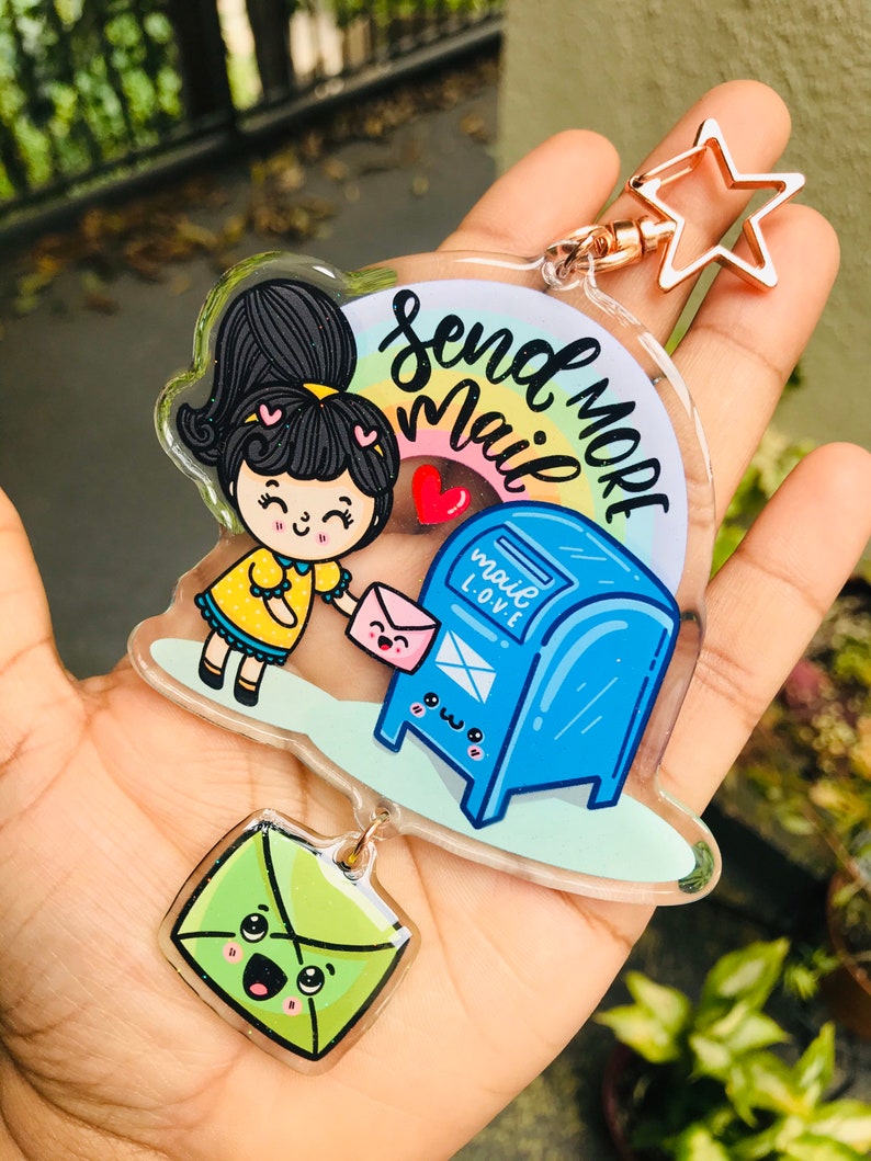 Pu&ograve; includere: Un portachiavi in acrilico colorato con una ragazza dei cartoni animati che tiene una busta rosa e una cassetta delle lettere blu con il testo "Send More Mail" e un arcobaleno. Il portachiavi include anche un ciondolo a forma di busta verde con un viso felice.