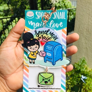 Pu&ograve; includere: Un portachiavi in acrilico colorato con una ragazza dei cartoni animati che tiene una busta rosa e una cassetta delle lettere blu con il testo "Send More Mail". Il portachiavi include anche una busta verde con un viso sorridente. Il testo "Spread Snail Mail Love" &egrave; stampato sulla carta dietro il portachiavi.