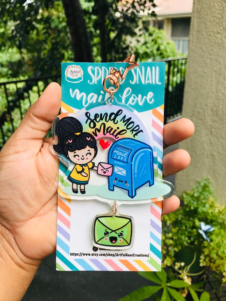 Pu&ograve; includere: Un portachiavi in acrilico colorato con una ragazza dei cartoni animati che tiene una busta rosa e una cassetta delle lettere blu con il testo "Send More Mail". Il portachiavi include anche una busta verde con un viso sorridente. Il testo "Spread Snail Mail Love" &egrave; stampato sulla carta dietro il portachiavi.