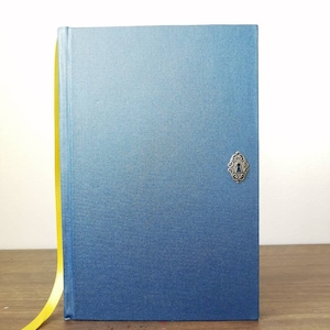 Puede incluir: Un cuaderno azul con un marcador de cinta amarilla y un dije de cerradura de plata.