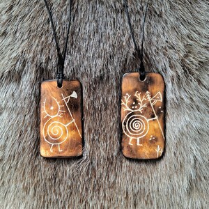 May include: Two wooden pendants with black cords, each featuring a different design. One pendant depicts a stylized figure with a spiral and a small axe, while the other shows a similar figure with antlers and a spiral. Both pendants have a brown wood background and white designs.
