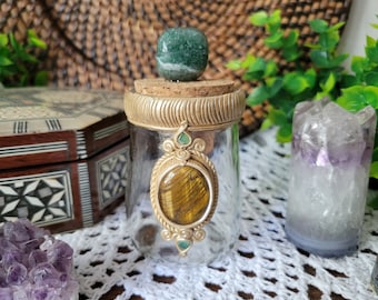 May include: A glass bottle with a gold lid and a cork stopper. The bottle is decorated with a tiger's eye stone and green gemstones. The bottle is sitting on a white crocheted surface.