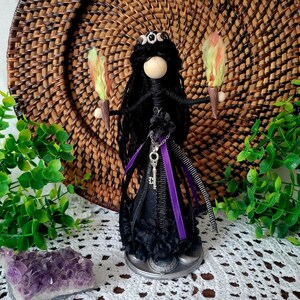 May include: A handcrafted doll with long black yarn hair, a smooth wooden bead head, and a black dress featuring purple ribbon accents. The doll holds two small torches with yellow and green flames and wears a silver-toned key pendant necklace.
