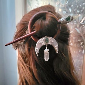 May include: A dark brown wooden hair fork featuring a circular ring and a pointed stick. A silver-toned crescent moon ornament with an embedded green stone and a dangling clear quartz crystal point is attached to the circular wooden hair accessory.
