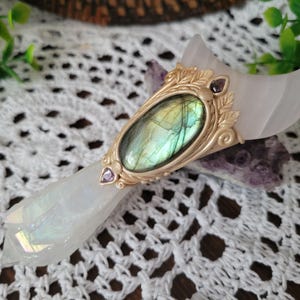 Peut inclure: Une baguette en quartz rose en forme de croissant avec un cabochon central en labradorite ovale serti dans une monture en métal doré ornée de feuilles. De petites pierres précieuses violettes sont incrustées en haut et en bas du sertissage.