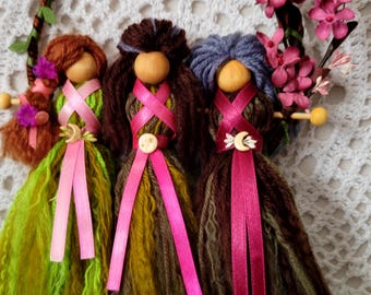 May include: A handcrafted wall hanging featuring three whimsical dolls with wooden heads and yarn hair in shades of brown, green, and blue. Each doll has a pink ribbon sash and a button or charm. The dolls are set against a white crocheted background, with a floral wreath.