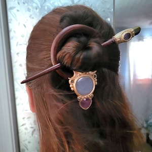 May include: A dark brown wooden hair fork featuring a circular ring and a pointed stick. The accessory is adorned with a gold-toned metal setting holding a large, smooth, pale blue oval stone and a smaller, faceted, deep purple teardrop-shaped stone.