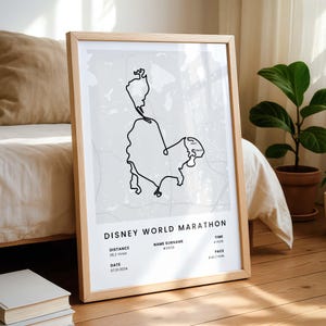 May include: A framed print featuring a black line drawing of the Disney World Marathon route on a gray background. The print includes details such as distance (26.2 miles), date (07.01.2024), and pace (9:43/mile). The frame is made of light-colored wood.