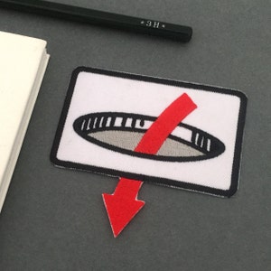 Puede incluir: Parche blanco con un círculo negro y gris con una línea diagonal roja que lo atraviesa. Una flecha roja apunta hacia abajo desde la parte inferior del parche.