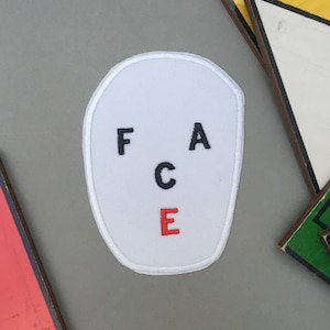 Puede incluir: Parche de tela blanco con la palabra "FACE" bordada en letras negras y rojas. Las letras están dispuestas en forma de diamante.