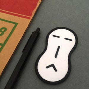 Puede incluir: Parche de fieltro blanco con un contorno negro en forma de cacahuete con una cara negra simple. La cara tiene dos líneas horizontales para los ojos, una línea vertical para la nariz y una línea curva para la boca.