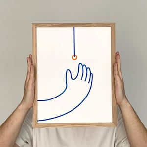 Puede incluir: Una persona sostiene un marco con una ilustración de una mano azul que alcanza un pequeño círculo naranja. El círculo está suspendido de una fina línea azul.