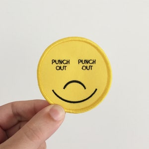Puede incluir: Parche bordado amarillo con una cara sonriente y el texto "Punch Out" a cada lado de los ojos.