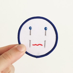 Puede incluir: Un parche blanco bordado con un borde circular azul. El parche presenta una cara triste con ojos azules y una boca roja.