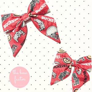 May include: Two red fabric hair bows with a repeating pattern of "Ohio State" and "Buckeyes" text, along with a football helmet and the Ohio State logo. The bows are on a white background with black polka dots.