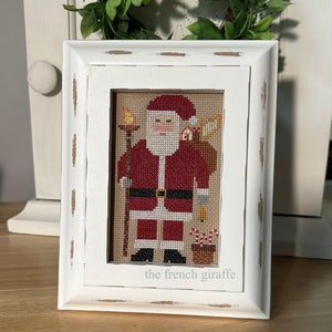 May include: A white framed cross-stitch of Santa Claus holding a sack of gifts and a lantern. The cross-stitch is signed "the french giraffe".