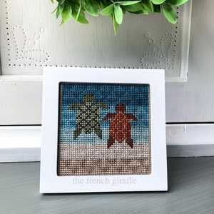 Puede incluir: Un punto de cruz enmarcado en blanco con un esquema de color azul, verde y naranja que representa dos tortugas marinas en una playa. El texto "the french giraffe" está en la parte inferior del punto de cruz.