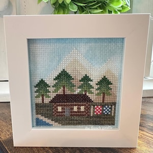 May include: A framed cross-stitch artwork depicting a brown cabin with a blue door and white windows, surrounded by green trees and snow-capped mountains. The sky is blue with white clouds. The artwork is signed "the french giraffe".