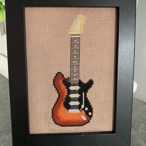 May include: A framed cross-stitch of a brown and black electric guitar with a cream-colored pickguard. The guitar is in a black frame with a beige mat.