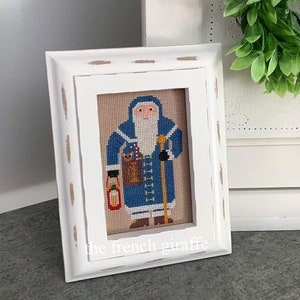 May include: A framed cross-stitch of Santa Claus in a blue coat, holding a lantern and a walking stick. The artwork is set against a tan background and framed in a distressed white wooden frame. The text "the french giraffe" is at the bottom.