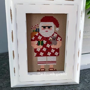 May include: A white framed cross-stitch of a Santa Claus wearing a red Hawaiian shirt with white flowers, sunglasses, and holding a gift and a drink. The cross-stitch is signed "the french giraffe".