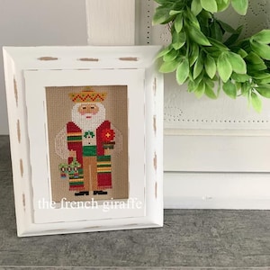 May include: A white framed cross-stitch of a Santa Claus wearing a sombrero and holding a bag of gifts. The cross-stitch is on a beige background and the frame has a distressed white finish. The text "the french giraffe" is visible at the bottom of the frame.