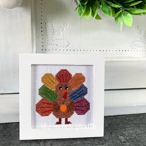 Op de afbeelding: Een ingelijst kruissteek van een kleurrijke kalkoen met een bruine pompoen. De kalkoen heeft rode, oranje, gele, groene, blauwe en paarse staartveren. Het kruissteek is in een wit frame met een witte passe-partout.
