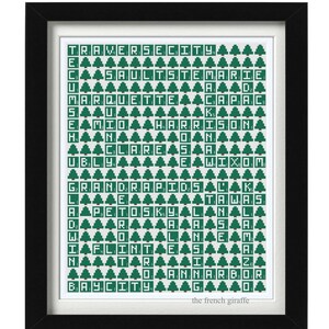 May include: Framed art print featuring a grid of green tiles spelling out Michigan city names, each row punctuated with a green tree icon. The print is set against a white background within a black frame, with the text "the french giraffe" at the bottom.