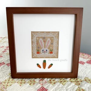 Op de afbeelding: Een ingelijst kruissteek van een wit konijn met oranje wortels. De lijst is van bruin hout en de passe-partout is wit. De tekst "the french giraffe" is onder de afbeelding gedrukt.