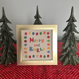 May include: A framed cross-stitch with a white background and colorful text that reads "Merry and Bright". The text is surrounded by a border of colorful circles. The frame is gold and the cross-stitch is sitting on a red and white polka dot surface.