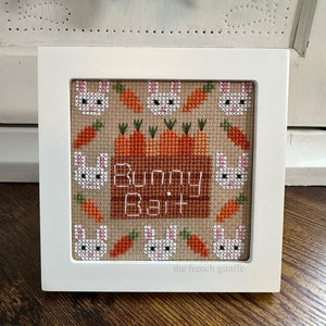 May include: A framed cross-stitch design featuring a brown wooden sign with the text "Bunny Bait" surrounded by white bunnies and orange carrots on a beige background.