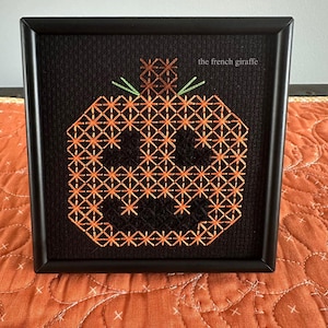 May include: Framed cross-stitch art featuring an orange pumpkin design on a black background. The pumpkin is stitched with orange thread, and the stem and leaves are green. The frame is black, and the text "the french giraffe" is visible.