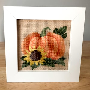 Könnte beinhalten: Ein gerahmtes Kreuzstich-Design mit einem Kürbis mit einer Sonnenblume und grünen Blättern. Der Hintergrund ist ein beigefarbener Stoff. Der Text "the french giraffe" ist in der rechten unteren Ecke sichtbar.