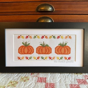 May include: A framed cross-stitch design featuring three orange pumpkins with green stems and leaves. The pumpkins are surrounded by a border of green, orange, and yellow leaves. The text "the french giraffe" is at the bottom of the design.