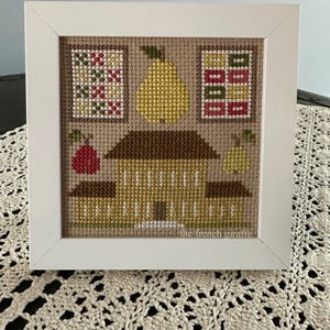 May include: A framed cross-stitch artwork depicting a house, pears, and geometric patterns in yellow, brown, red, and green. The artwork is set against a tan background and framed in a white square frame. The text "the french giraffe" is visible.