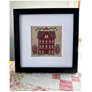 May include: A framed cross-stitch of a red house with white windows and a green roof. The house is surrounded by green bushes with pink flowers. The text "The French Giraffe" is visible in the bottom right corner of the image.