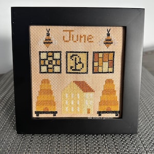 May include: A framed cross-stitch design featuring the word "June" and bee motifs. The design includes a beehive, a house, and a letter "B" in a yellow and brown color scheme.