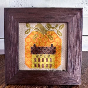 May include: A framed cross-stitch design of a pumpkin with the year 2022 stitched in the center. The pumpkin is orange and yellow with a brown stem and green leaves. The frame is dark brown wood.