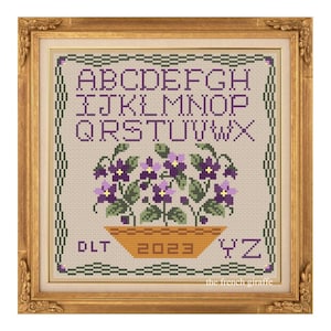 May include: Cross-stitch sampler in a gold frame. The design features the alphabet, a basket of purple flowers, and the year 2023. The text "the french giraffe" is in the bottom right corner. The frame is rectangular.