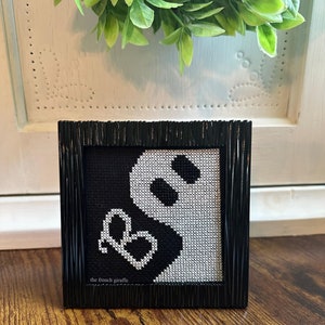 May include: Black and white cross-stitch embroidery of a ghost with a heart in a black frame. The embroidery is signed "the french giraffe".