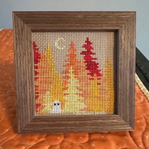 Op de afbeelding: Een ingelijste kruissteeklandschap met een boslandschap met bomen in tinten geel, oranje en rood. Een halve maan is in de lucht gestikt. Het frame is van hout.