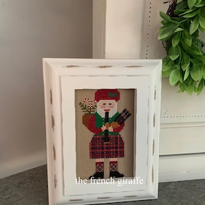 Könnte beinhalten: Ein weiß gerahmtes Kreuzstichbild eines Weihnachtsmanns, der Dudelsack spielt. Der Weihnachtsmann trägt ein rot-grünes Outfit mit einem karierten Kilt. Der Rahmen hat eine weiße, abgenutzte Oberfläche. Der Text "the french giraffe" befindet sich am unteren Rand des Rahmens.