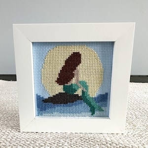 May include: A framed cross-stitch picture depicting a mermaid silhouette against a yellow moon and blue water. The mermaid has brown hair and a green tail. The artwork is in a white square frame, suitable for wall decor.