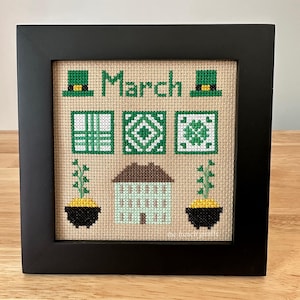 May include: A framed cross-stitch design featuring a green and white house, shamrocks, and leprechaun hats. The design is titled "March".